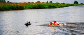 jeden strażak płynie po wodzie w pomarańczowym ubraniu do drugiego w czarnym w oddali zarośla
