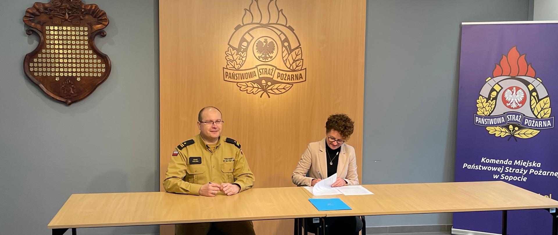 Przy stole siedzi Magdalena Czarzyńska-Jachim pełniąca funkcje prezydenta Miasta Sopotu która podpisuje umowe oraz mt. bryg. Adam Halasa po komendanta miejskiego w Sopocie . W tle loga PSP