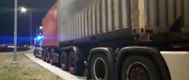 Na pierwszym planie: bok przeładowanego, wieloosiowego zespołu pojazdów z dwoma kontenerami. W tle: za nienormatywną ciężarówką stoi oznakowany furgon mazowieckiej Inspekcji Transportu Drogowego z włączonymi niebieskimi światłami.