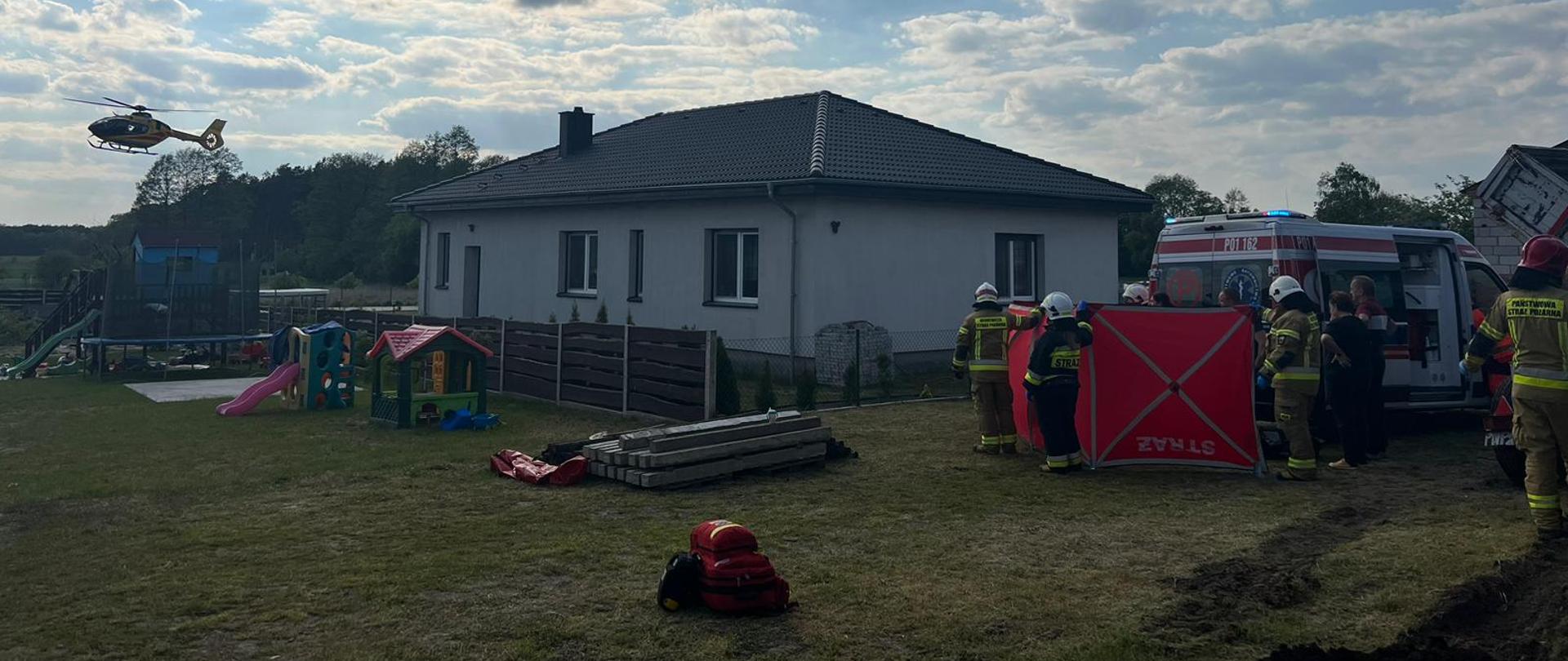 Na zdjęciu karetka ZRM wraz ze strażakami i policjantami, w tle startujący helikopter LPR
