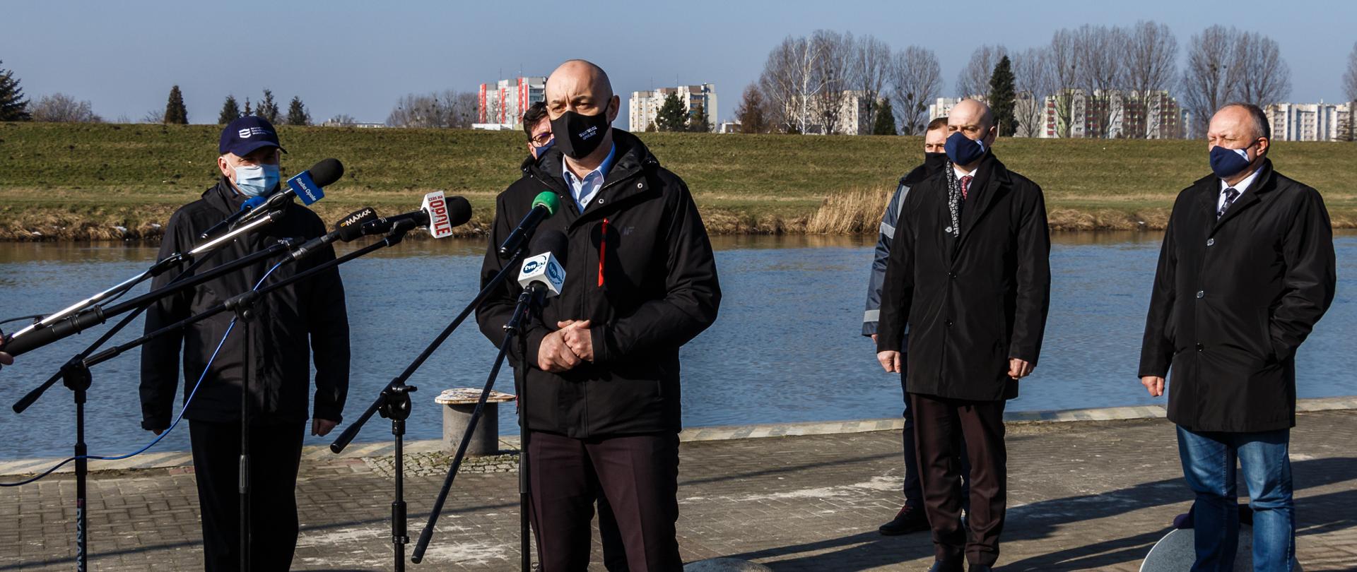 Konferencja prasowa wojewody opolskiego i PGW WP. Na zdjęciu stoi 5 meżczyzn na tle rzeki Odry. Na środku kadru wojewoda opolski przemawia do zgromadzonych dziennikarzy, mówi przez mikrofon. Wojewoda jest ubrany w ciemny garnitur, na twarzy ma ciemną maseczkę ochronną. Na dalszym planie pozostali mężczyźni. w dalekim tle widać panoramę Opola - tj. wieżowce. 