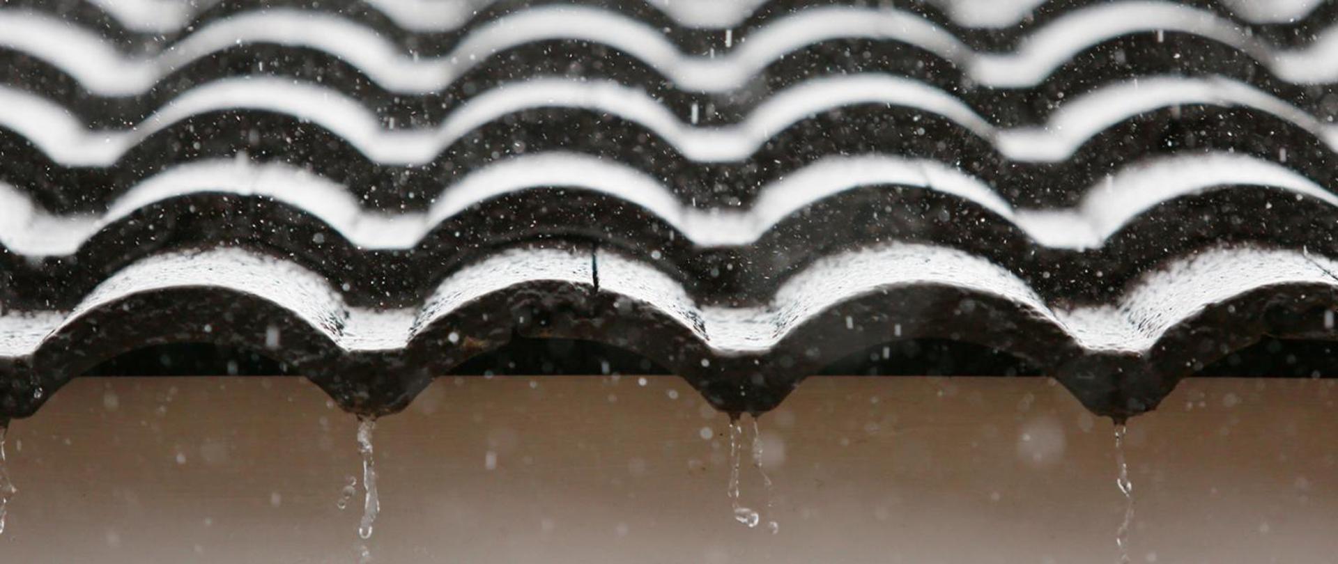 Na zdjęciu widać część falistego dachu z ciemnych dachówek, po którym spływa deszcz.