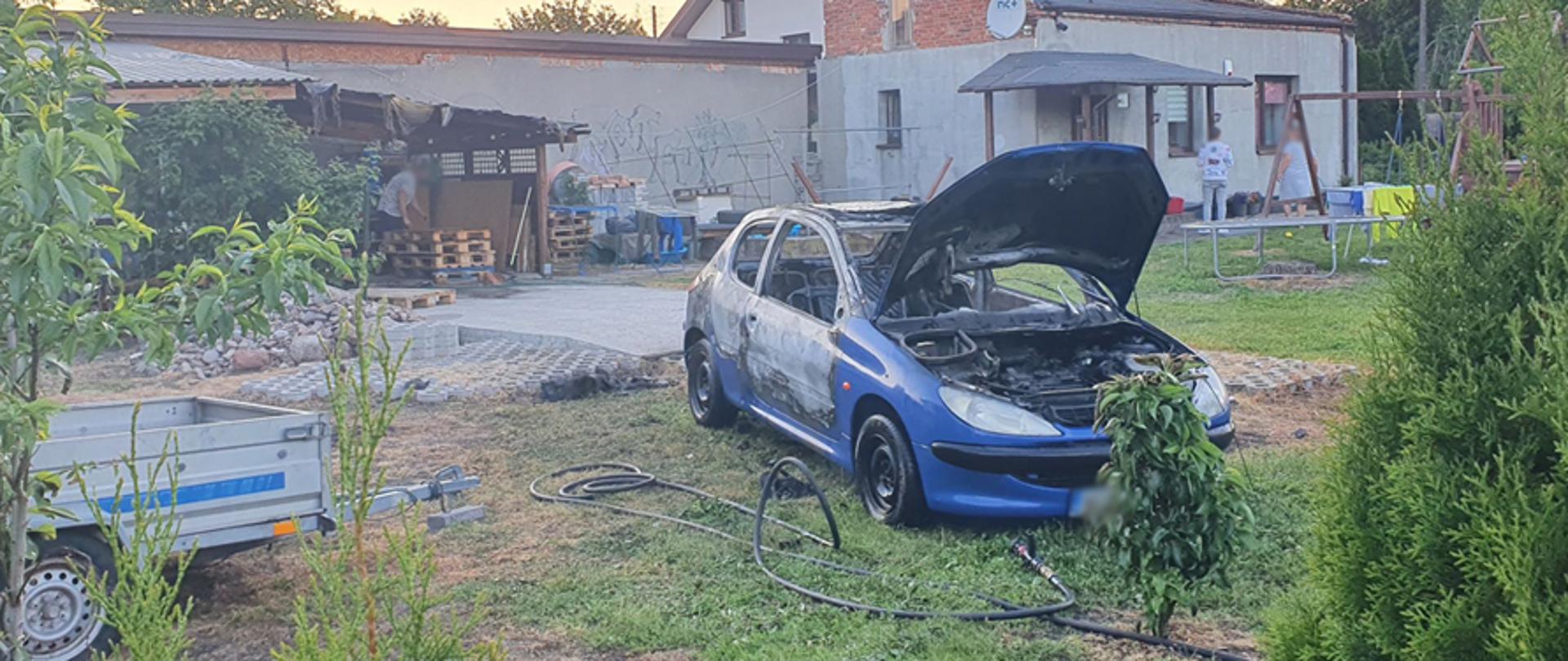 Na zdjęciu widoczny samochód koloru niebieskiego stojący na prywatnej posesji. Widoczne spalone wnętrze pojazdu oraz rozwiniętą linię szybkiego natarcia z samochodu strażackiego.