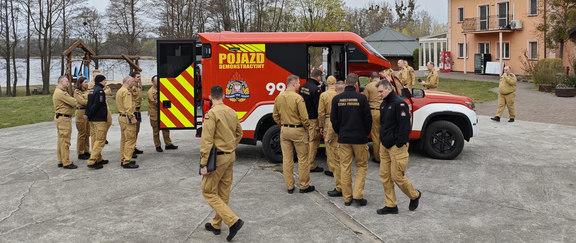 Pojazd demonstracyjny, w kolorze czerwonym, stoi na środku placu. Wokół pojazdu jest zgromadzonych kilkudziesięciu strażaków. Tylne drzwi pojazdu, oklejone w żółte pasy, są otwarte.