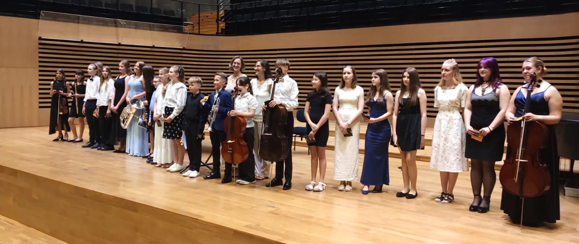 Około dwadzieścioro uczniów stoi w rzędzie ze swoimi instrumentami na scenie, za nimi stoją dwie kobiety.
