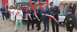 na zdjęciu st. bryg. Dariusz Gieroń - Zastępca Opolskiego Komendanta Wojewódzkiego Państwowej Straży Pożarnej podczas przecinania wstęgi w tle nowy samochód ratowniczo -gaśniczy.