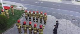 Zdjęcie przedstawia dwie zmiany służbowe stojące naprzeciw siebie podczas uroczystego podniesienia flagi na maszt, gdzie przy maszcie stoi trzech strażaków podnoszących flagę. Uroczystość ma miejsce na placu zewnętrznym Komendy Powiatowej Państwowej Straży Pożarnej w Krośnie Odrzańskim.