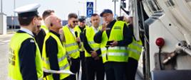 Grupa osób w kamizelkach odblaskowych stoi obok ciężarówki, uczestnicząc w szkoleniu z zakresu kontroli transportu drogowego.