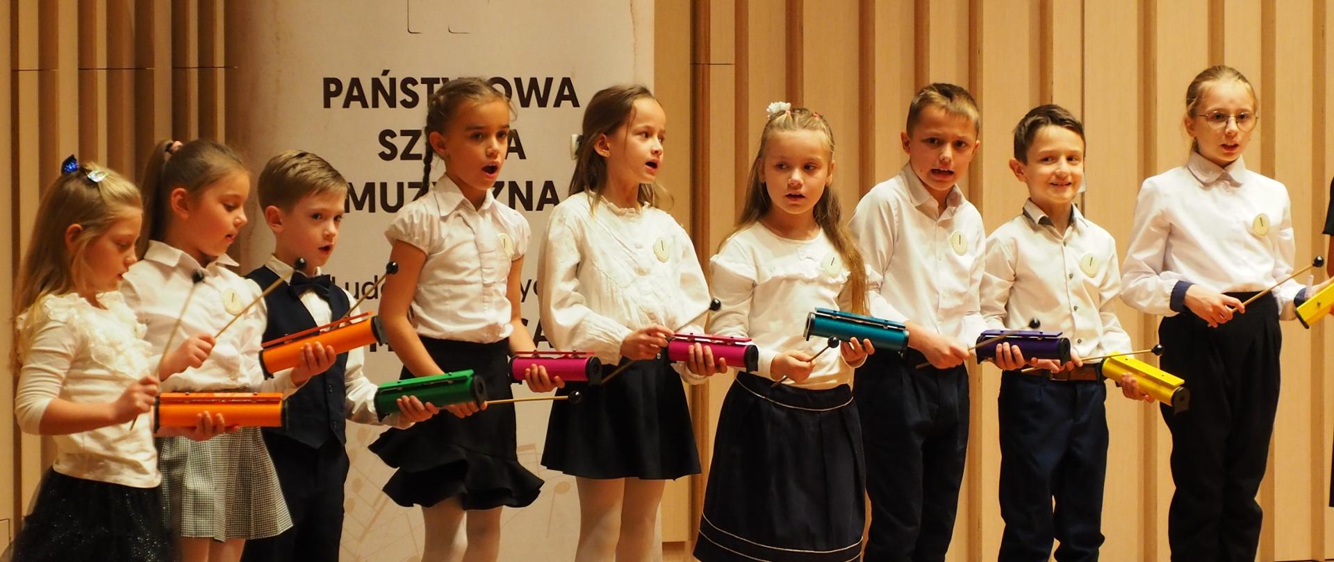 Uczniowie z instrumentami perkusyjnymi na scenie w sali koncertowej.