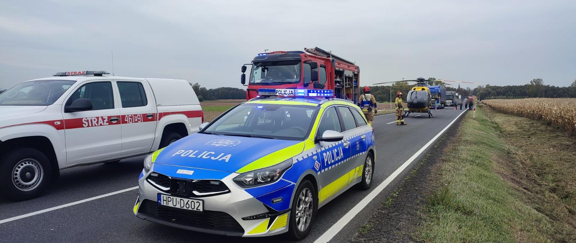 Na drodze wiejskiej stoją pojazdy służb ratunkowych — radiowóz policji, wozy straży pożarnej i śmigłowiec Lotniczego Pogotowia Ratunkowego. Policjanci i strażacy zabezpieczają miejsce zdarzenia. Droga jest zablokowana, a w tle widać pola i las.