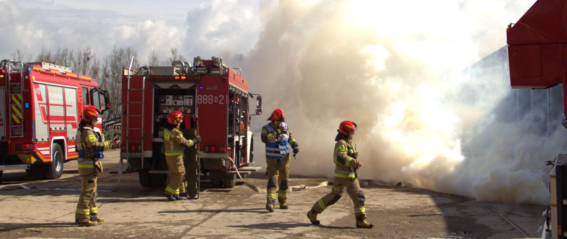 Dwa wozy strażackie z lewej strony. Przy nich strażacy. Z prawej dużo białego dymu.