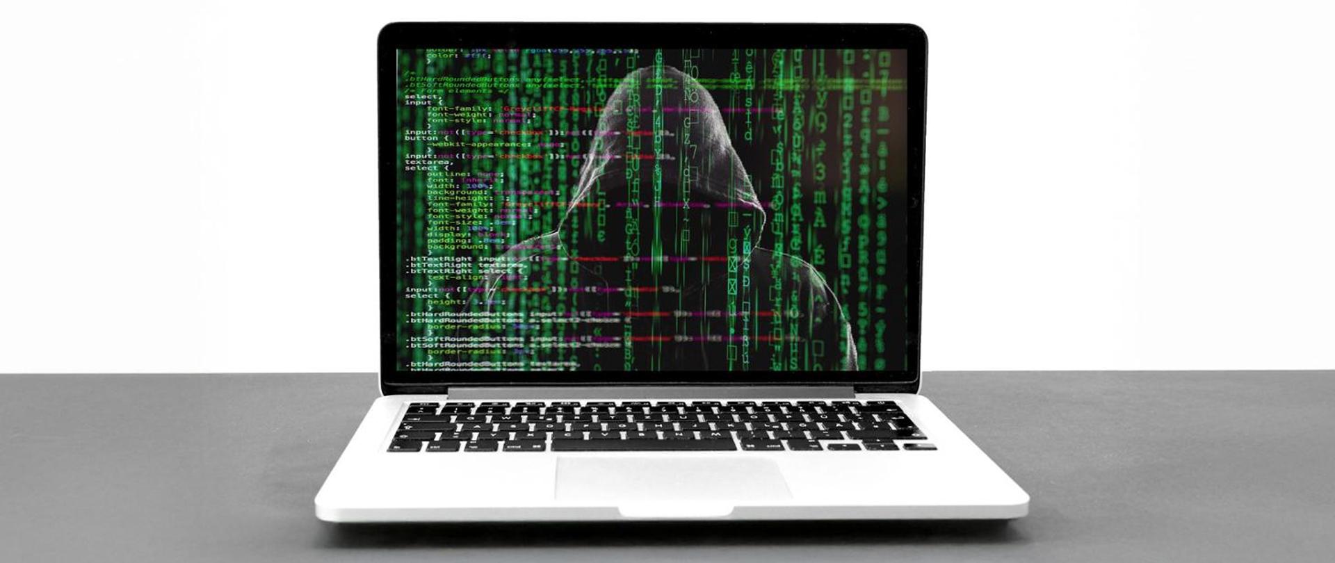 Laptop, na jego ekranie zakapturzona postać, w tle kody.