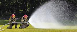 Na polanie dwóch strażaków w pozycji klęczącej trzyma waz strażacki i podaje przed siebie rozproszony prąd wody. Strażacy mają piaskowe mundury i czerwone hełmy. W tle widać wysokie drzewa.