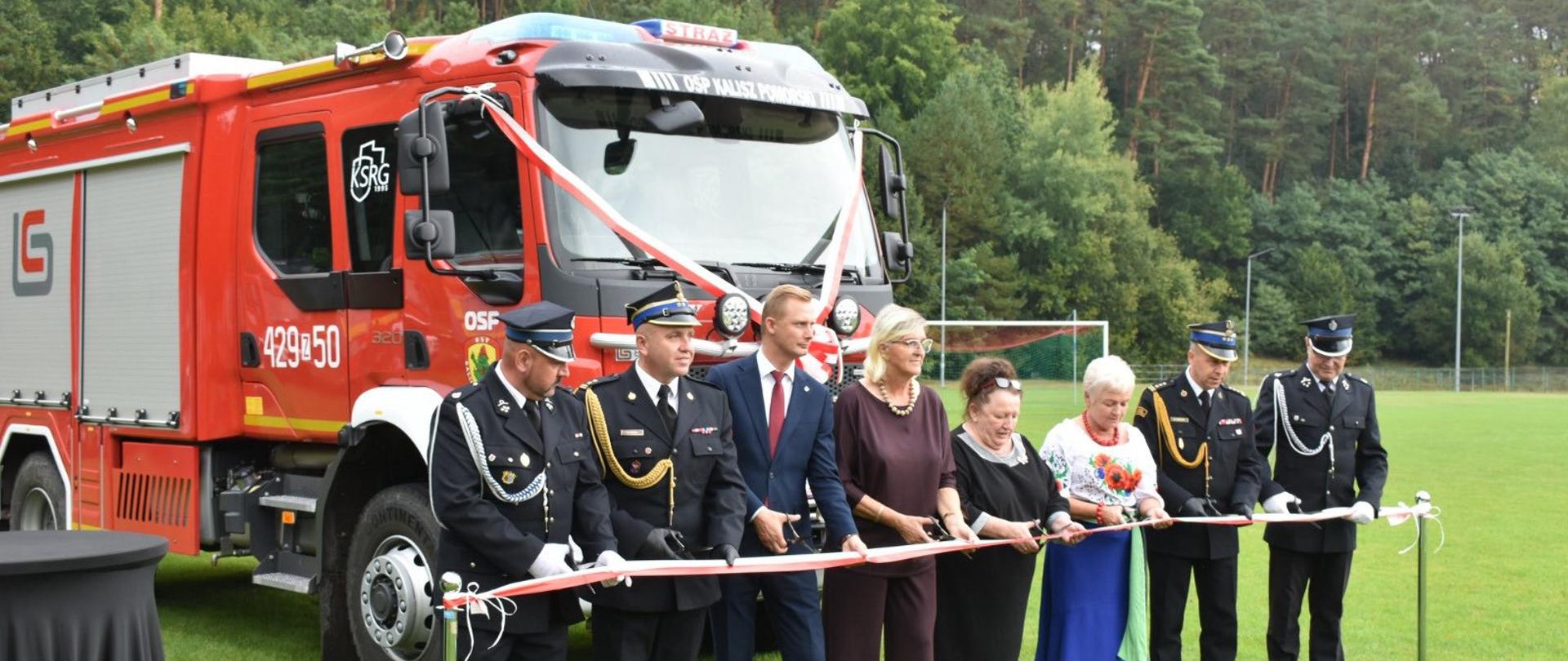 Zdjęcie przedstawia grupę dziewięciu osób stojących w rzędzie na trawiastym polu. Znajdują się przed dużym, czerwonym wozem strażackim.
Wszyscy trzymają długą, biało-czerwoną wstęgę, którą wspólnie przecinają za pomocą nożyczek. Ubrani są w różne stroje: niektórzy mają formalne mundury, a inni eleganckie ubrania. W tle widać las i fragment boiska sportowego z bramką.
