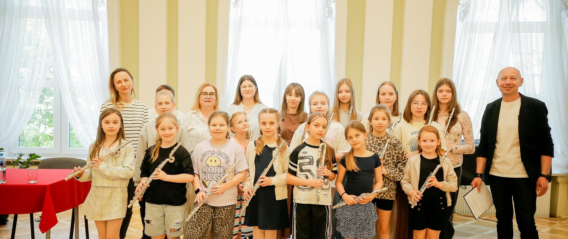 Zdjęcie grupowe przedstawiające kilkanaście osób, głównie dzieci i młodzież, pozujących na niewielkim podwyższeniu lub scenie w eleganckiej, klasycznej sali. W pierwszym rzędzie stoi siedem dziewczynek w wieku szkolnym, z których każda trzyma w dłoniach flet poprzeczny. Dziewczynki są ubrane w swobodne, letnie stroje – sukienki, spódniczki, spodenki oraz koszulki.
Za nimi, w drugim rzędzie, stoi grupa starszych dziewcząt oraz kilka dorosłych kobiet, prawdopodobnie nauczycielek lub opiekunek. Po prawej stronie grupy stoi dorosły, łysy mężczyzna w czarnej marynarce, białym tiszercie i ciemnych spodniach, uśmiechający się do aparatu i trzymający w ręku dyplom lub dokumenty.
Tło stanowią duże, wysokie, łukowate okna z białymi, delikatnymi firankami, rozdzielone dekoracyjnymi kolumnami ze złotymi akcentami. Po lewej stronie widoczny jest fragment stołu przykrytego czerwonym obrusem. Całe pomieszczenie ma jasne, pastelowe ściany i drewniany parkiet ukierunkowany w jodełkę. Wszyscy uczestnicy spotkania patrzą w stronę obiektywu.