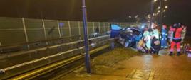 Na zdjęciu widać uszkodzony pojazd. Na pierwszym planie widać chodnik, barierki drogowe i asfalt. Pogoda deszczowa. Pora Nocna. W oddali funkcjonariusze PSP oraz ratownicy PRM. Trwa akcja ratunkowa.