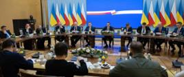 Szerokie ujęcie. Przedstawiciele delegacji polskiej i ukraińskiej siedzą przy okrągłym stole. W tle ekran i flagi. 