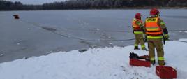Na zdjęciu widać strażaka na saniach lodowych znajdującego się na zamarzniętym akwenie w asekuracji strażaków będących na brzegu.