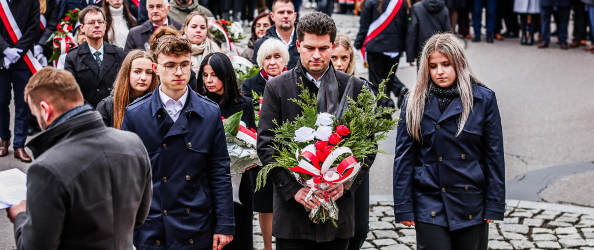 Nauczyciel z uczniami składa wiązankę biało-czerwonych kwiatów pod Pomnikiem Wolności w Mielcu podczas obchodów Narodowego Święta Niepodległości, w otoczeniu uczestników uroczystości.