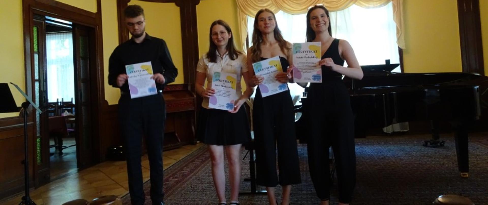 Zdjęcie kolorowe. Czworo absolwentów w strojach galowych trzymających dyplomy. W tle fortepian.