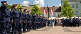 Zdjęcie przedstawia uroczystość Święta Policji w Rybniku, odbywającą się na brukowanym placu miejskim w słoneczny dzień. W centralnej części kadru, po lewej stronie, stoi rząd policjantów w ciemnogranatowych mundurach i granatowych czapkach. Są oni ustawieni w szeregu, zwróceni w prawo, a ich postawa sugeruje dyscyplinę i powagę. Pierwszy policjant z lewej strony unosi rękę do czoła, salutując. W tle, po prawej stronie zdjęcia, widać maszt z powiewającą biało-czerwoną flagą Polski. Za masztem widoczna jest grupa ludzi zgromadzonych, niektórzy z nich również ubrani są w mundury, inni w stroje cywilne. Poza grupami ludzi, w tle zdjęcia widać budynki, kamienice, z jasnymi elewacjami i zielonymi drzewami.