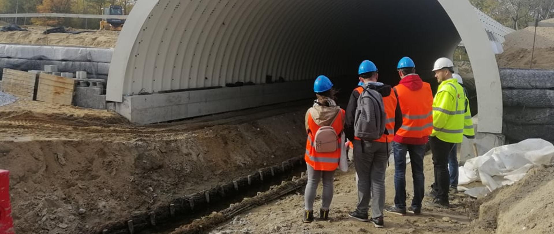 Ludzie w kaskach i kamizelkach zwróceni tyłem. Przed nimi tunel. wokół plac budowy, na ziemi piasek. 