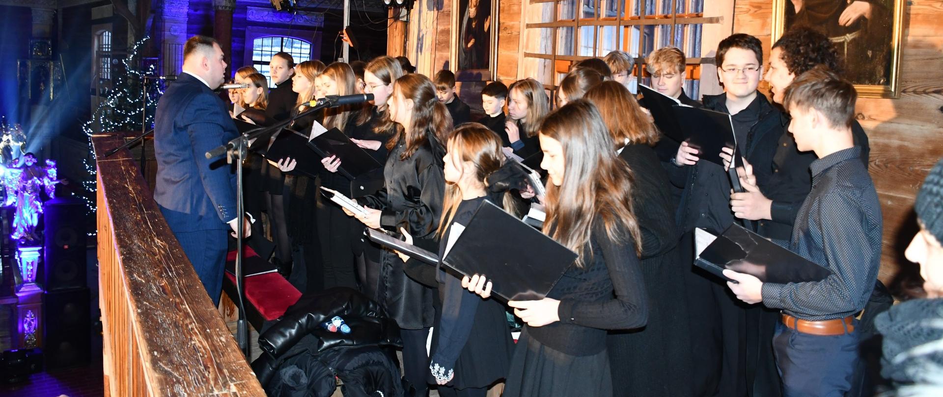 Występ chóru podczas koncertu w drewnianym, zabytkowym kościółku. Chórzyści ubrani są na czarno i trzymają w rękach czarne teczki z nutami. Śpiewacy ustawieni są na chórze w dwuszeregu. Przed chórem stoi dyrygent. Na zdjęciu oprócz chóru wyeksponowane są drewniane elementy konstrukcyjne kościółka, obrazy świętych i niebieskie oświetlenie. 