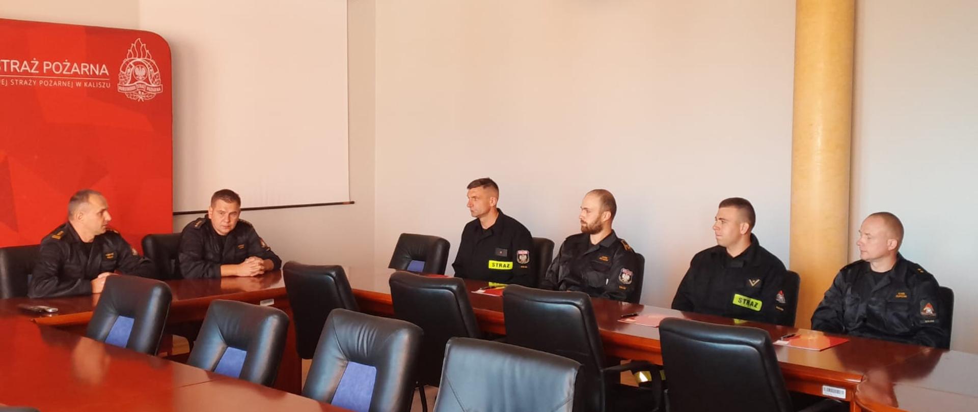 Widać umundurowanych funkcjonariuszy. Umundurowanie w kolorze czarnym. Światło naturalne. Pomieszczenie wewnątrz. Drewniany stół, brązowy oraz ekran wyświetlania.