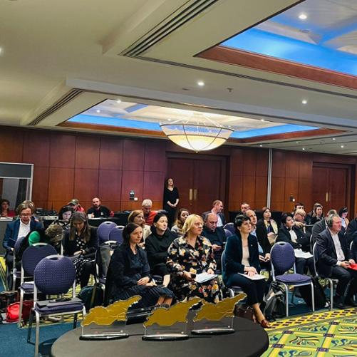 Grupa osób siedzi w 4 rzędach na sali konferencyjnej i słucha przemówienia