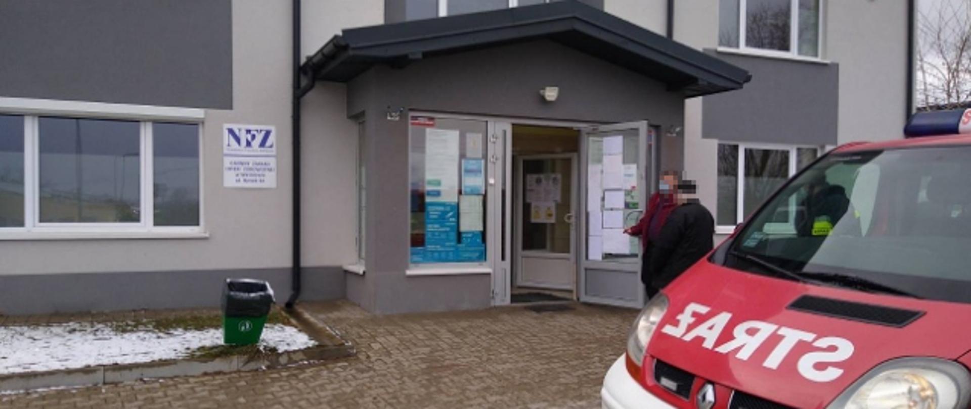 Przed przychodnią stoi zaparkowany samochód pożarniczy, który dowozi pacjentów na szczepienia. 