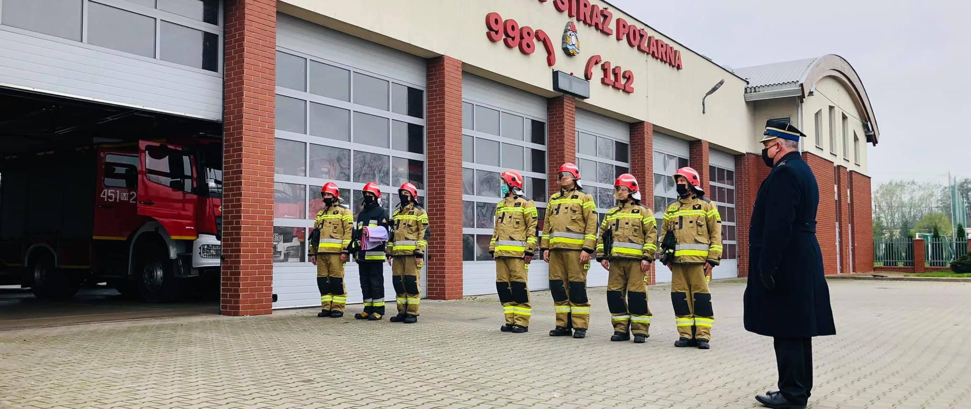 Zdjęcie przedstawia apel zorganizowany z okazji 11 listopada. Widoczna jest cała zmiana służbowa składająca się z 7 strażaków ubranych w pełne umundurowanie bojowe typu Nomexu wraz z hełmami strażackimi. Na zdjeciu znajduje się również komendant powiatowy PSP ubrany w ubranie wyjściowe. W tle znajdują się garaże z pojazdami pożarniczymi. 