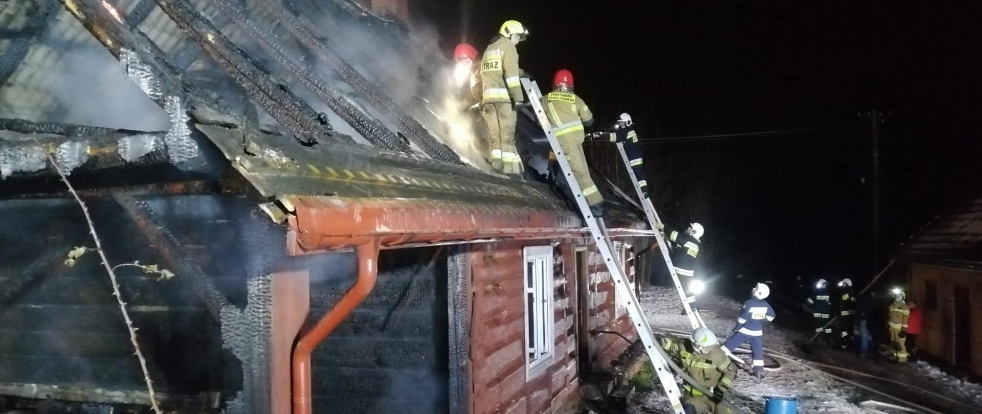 Kolorowa fotografia przedstawia nocną akcję gaśniczą straży pożarnej gaszenia brązowego, drewnianego budynku mieszkalno-gospodarczego. Teren jest pokryty śniegiem, z budynku wydobywa nieduża ilość dymu a teren akcji jest oświetlony przez strażaków. Na pierwszym planie widoczny spalony narożnik budynku. W głębi kilku strażaków w ubraniach bojowych, w hełmach na głowach pomaga gasi pożar przy użyciu dwóch drabin pożarniczych a kilku pozostałych w pobliżu budynku pomagają w akcji gaśniczej. Przed częściowo spalonym budynkiem widoczne białe węże pożarnicze wykorzystywane go podawania wody na pożar.