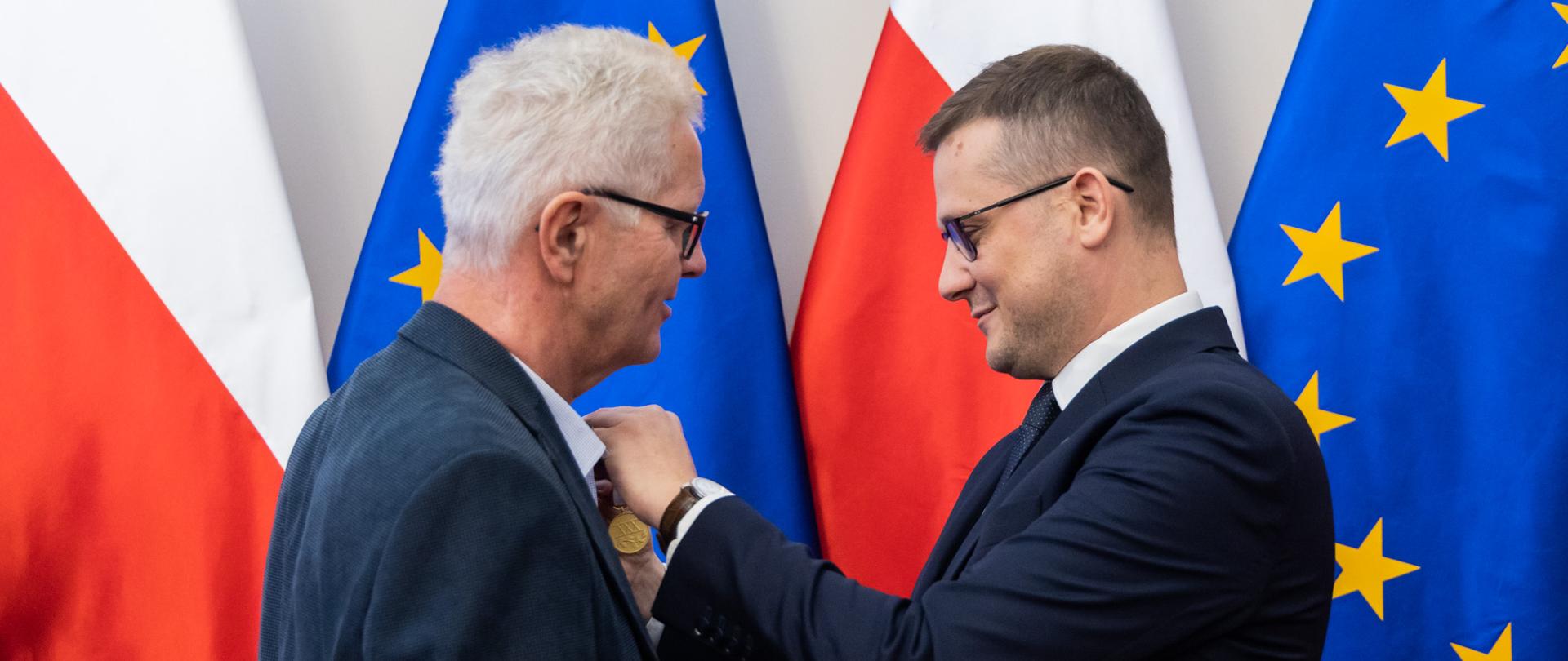 Wojewoda Kujawsko-Pomorski Michał Sztybel przypina złoty medal do marynarki starszego mężczyzny. Obaj mężczyźni noszą okulary i patrzą na wpinane odznaczenie. W tle znajdują się flagi Polski i Unii Europejskiej. 