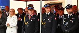Na trybunie honorowej pod czerwonym namiotem stoją zaproszeni goście mundurowi ubrani w mundury osoby cywilne w garnitury. Przed nimi biało-czerwone godło Polski z tyłu widoczny budynek ratusza