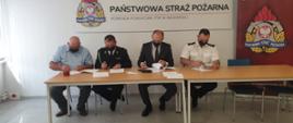 4 mężczyzn siedzących za stołem, podpisuje kartki z porozumieniem. Dwóch z nich jest ubranych w mundury, jeden z nich w ciemnym garniturze, czwarty w jasnobłękitnej koszuli. Za nimi dwie plansze z napisami "Państwowa Straż Pożarna" z logiem Państwowej Straży Pożarnej.