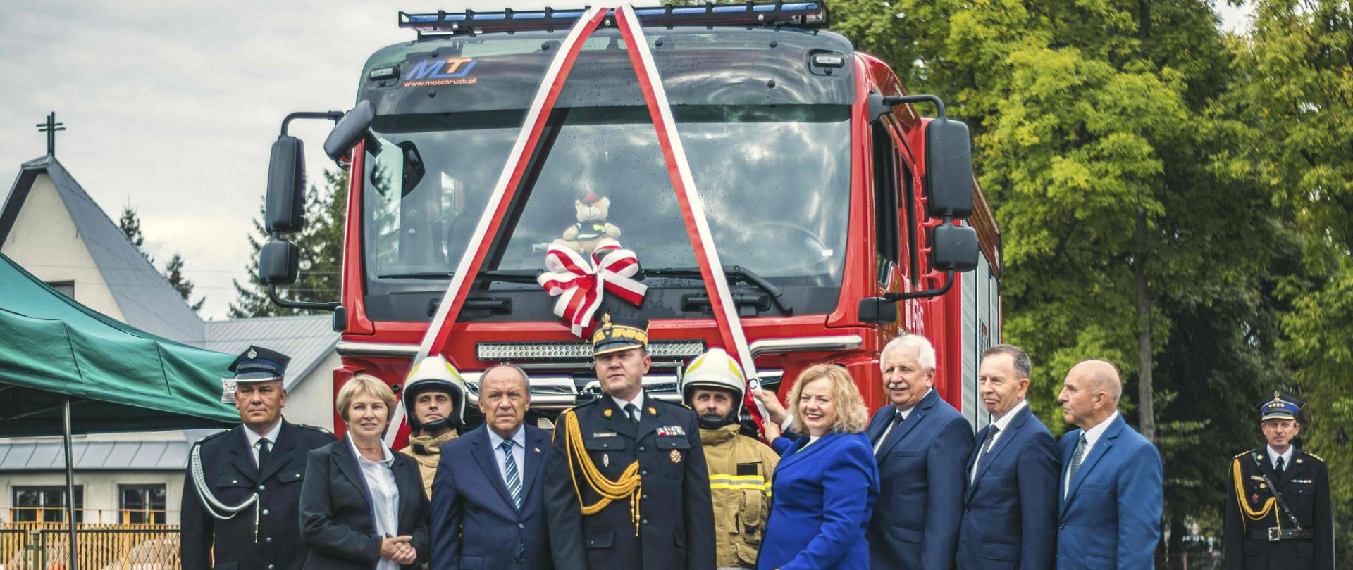 Przed przodem samochodu strażackiego, który przez szybę ma przewiązaną biało czerwoną wstęgę i kokardę stoi 9 osób: 4 mężczyzn w garniturach oraz 2 kobiety, 1 strażak PSP w mundurze wyjściowym oraz 2 OSP w ubraniach specjalnych. Po prawej stronie z boku stoi 1 strażak PSP w mundurze wyjściowym. W tle namiot, budynek oraz drzewa.