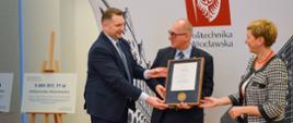 Minister Czarnek stoi i podaje mężczyźnie w garniturze dokument w ramkach, obok stoi kobieta w bluzce w biało-czarną kratkę, za nimi ścianka z napisem Politechnika Wrocławska.