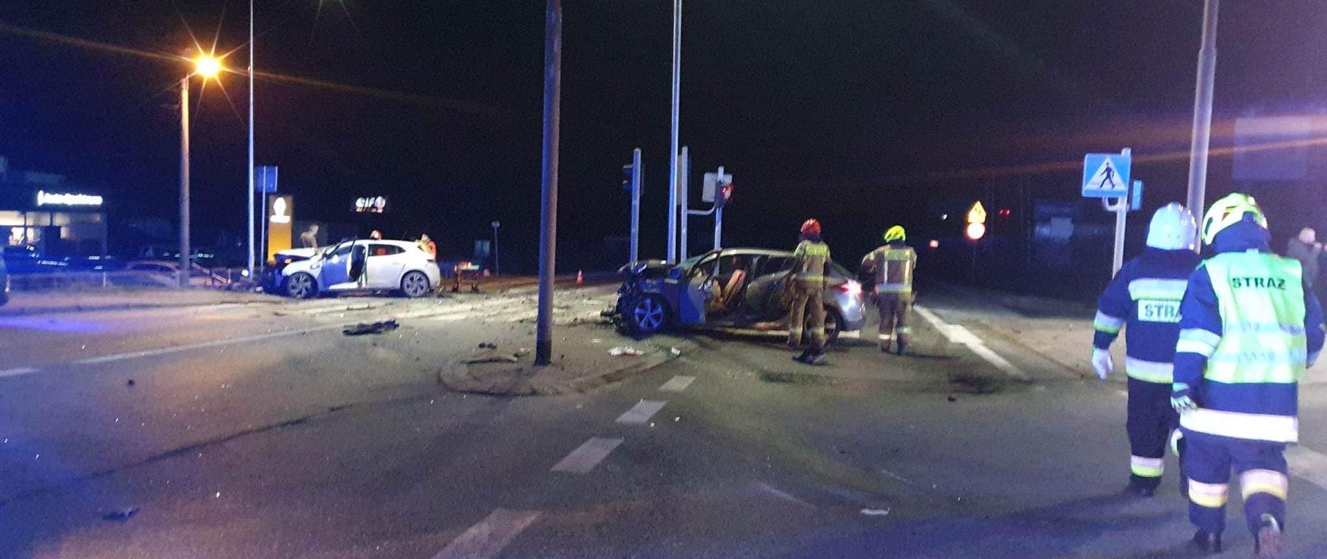 Na zdjęciu dwa pojazdy osobowe po zderzeniu znajdujące się na skrzyżowaniu. Obok pojazdów strażacy w ubraniach specjalnych.