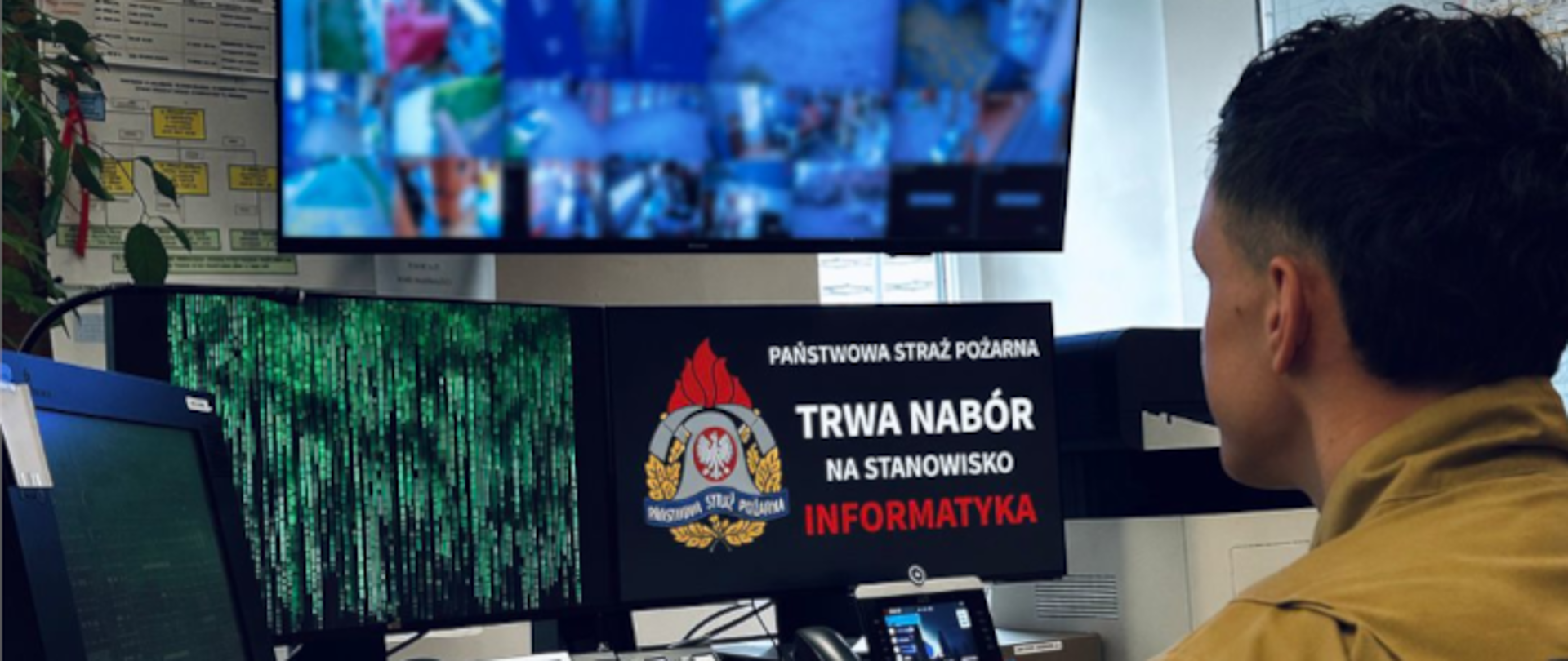 Strażak siedzi przy stanowisku komputerowym i patrzy na kilka monitorów. Na jednym z ekranów widać podgląd z kamer monitoringu, a na innym grafikę z napisem po polsku: „Państwowa Straż Pożarna – trwa nabór na stanowisko informatyka”.