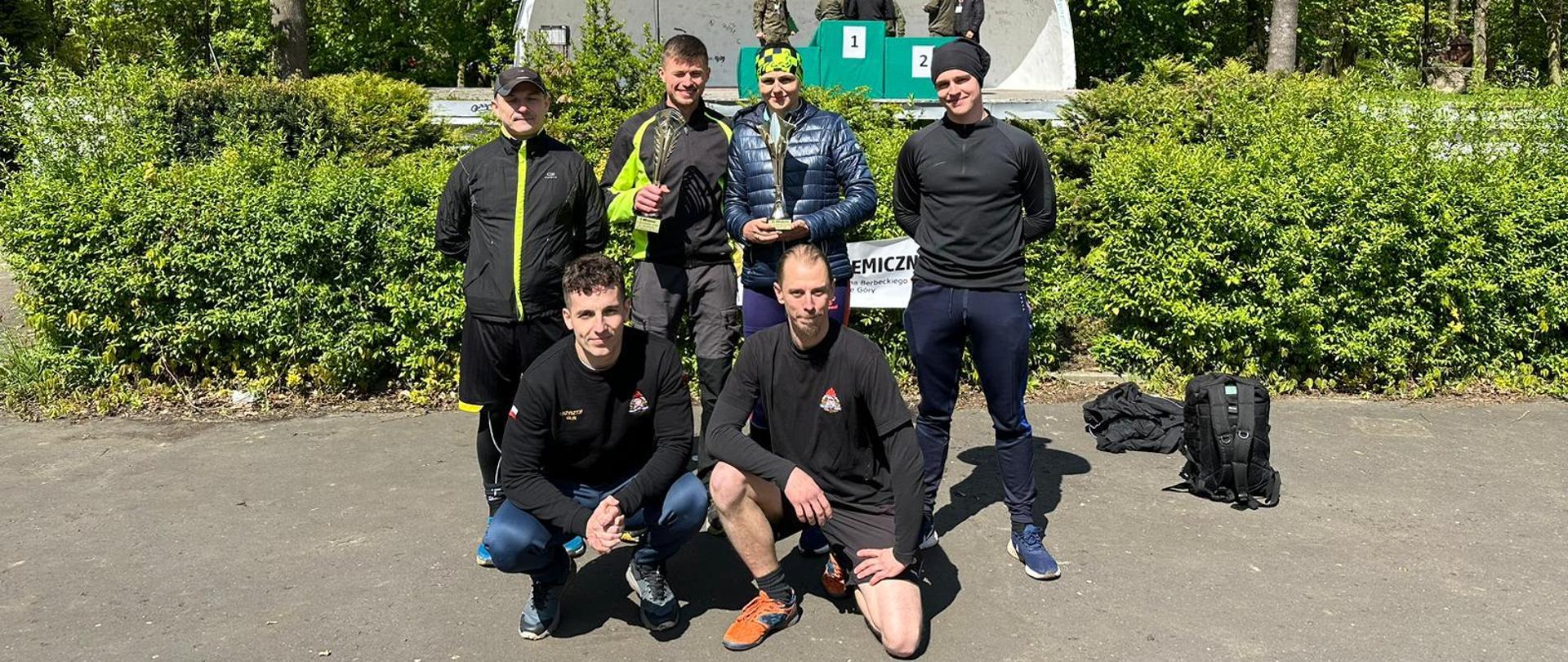 Na zdjęciu 6 osób. W pierwszym rzędzie kuca dwóch mężczyzn w drugim stoi trzech mężczyzn i jedna kobieta. Kobieta i mężczyzna stojący po jej lewej stronie trzymają w rękach puchar.