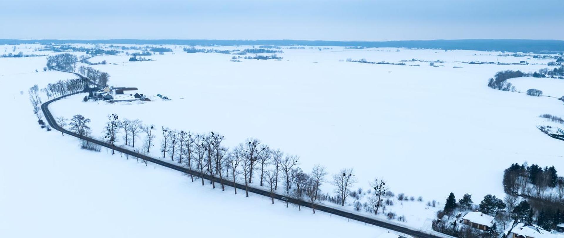 Zimowy, szeroki krajobraz sfotografowany z lotu ptaka. Przez śnieżne, białe pola prowadzi długa, kręta droga obsadzona rzędami drzew bez liści. Po bokach drogi widać rozproszone zabudowania i niewielkie skupiska drzew. Cała okolica jest pokryta jednolitą warstwą śniegu, a nad horyzontem unosi się jasne, zachmurzone niebo.