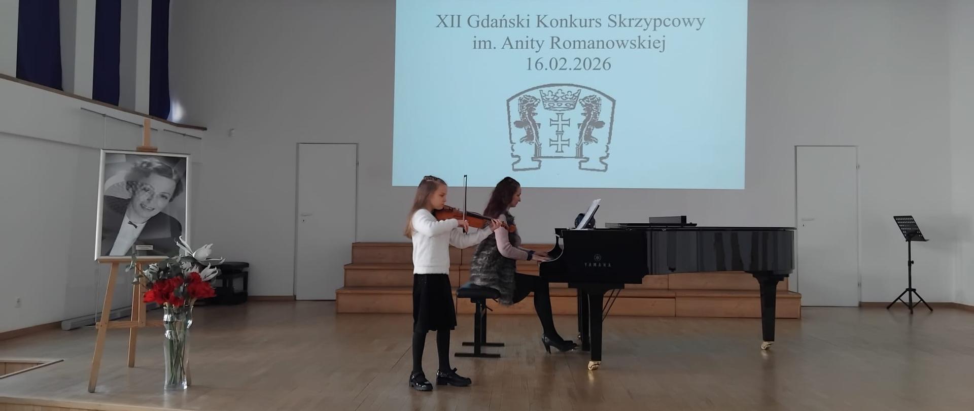 Na scenie sali koncertowej stoi dziewczynka grająca na skrzypcach. Ma na sobie białą bluzkę, czarną spódnicę, czarne rajstopy i czarne buty. Obok niej, po prawej stronie, przy fortepianie marki Yamaha siedzi akompaniatorka i gra z nut ustawionych na pulpicie. W tle na ścianie wyświetlony jest slajd z napisem: „XII Gdański Konkurs Skrzypcowy im. Anity Romanowskiej 16.02.2026” oraz herbem Gdańska. Sala jest jasna, z drewnianą podłogą i prostym, nowoczesnym sufitem.