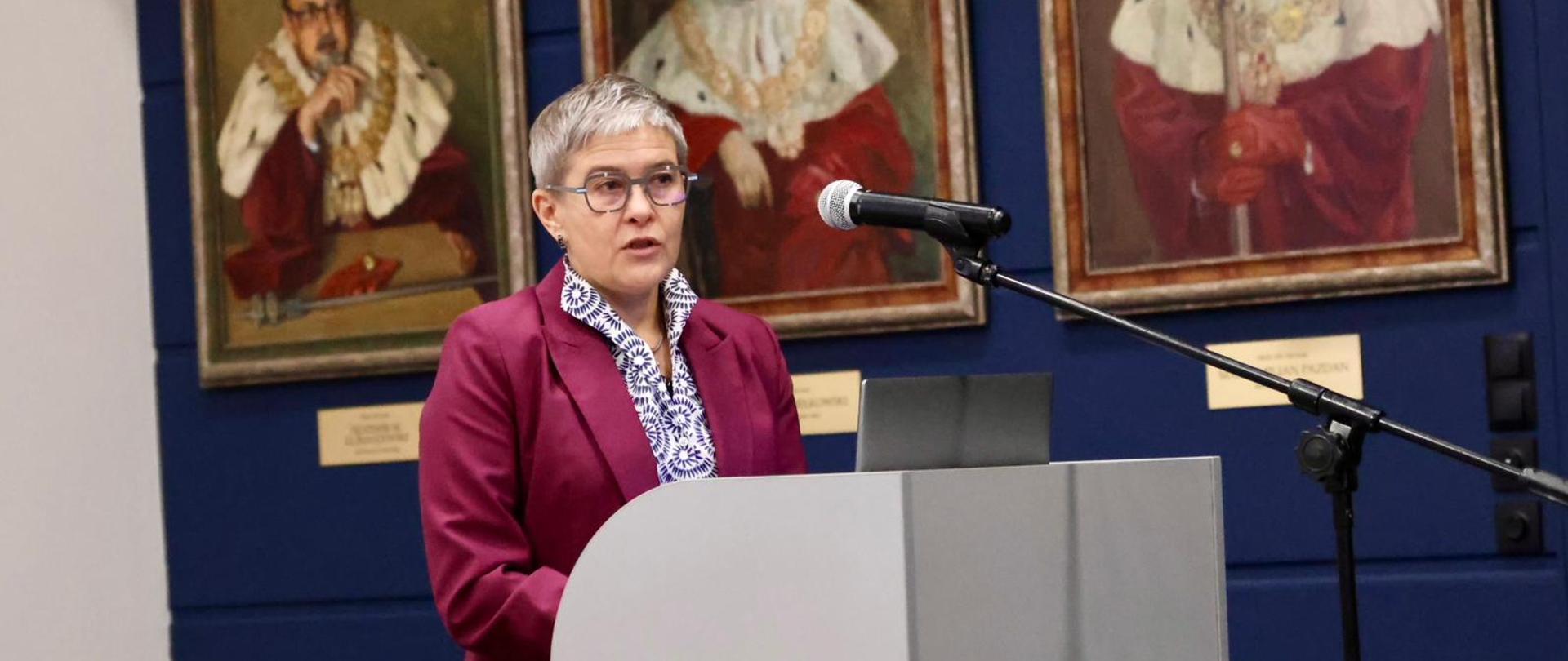 Wiceminister Mrówczyńska stoi za mównicą i mówi do mikrofonu na stojaku, za nią na ścianie wiszą trzy portrety mężczyzn w czerwonych ceremonialnych strojach.