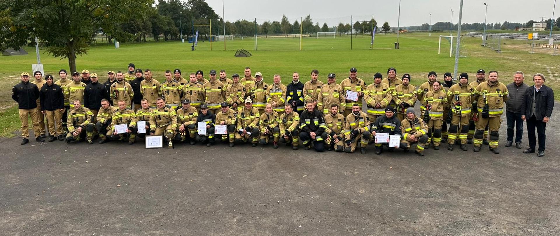 Na zdjęciu widoczni strażacy z dyplomami, po zakończonych zawodach. W tle duża grupa pozostałych strażaków. Na dalszym planie widoczne boisko sportowe.