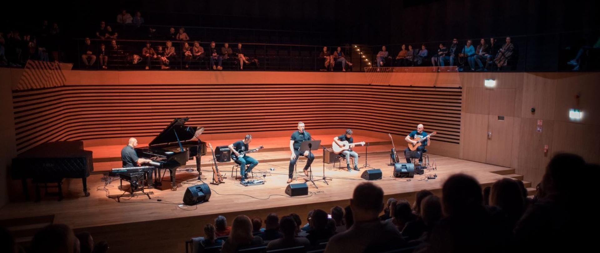 Pięciu mężczyzn na scenie sali koncertowej PSM w Jastrzębiu-Zdroju, po lewej stronie pianista gra na fortepianie, jeden mężczyzna siedząc śpiewa do mikrofonu, trzech gra na gitarach. Z przodu widoczna tyłem siedząca publiczność, nad sceną również siedząca publiczność.