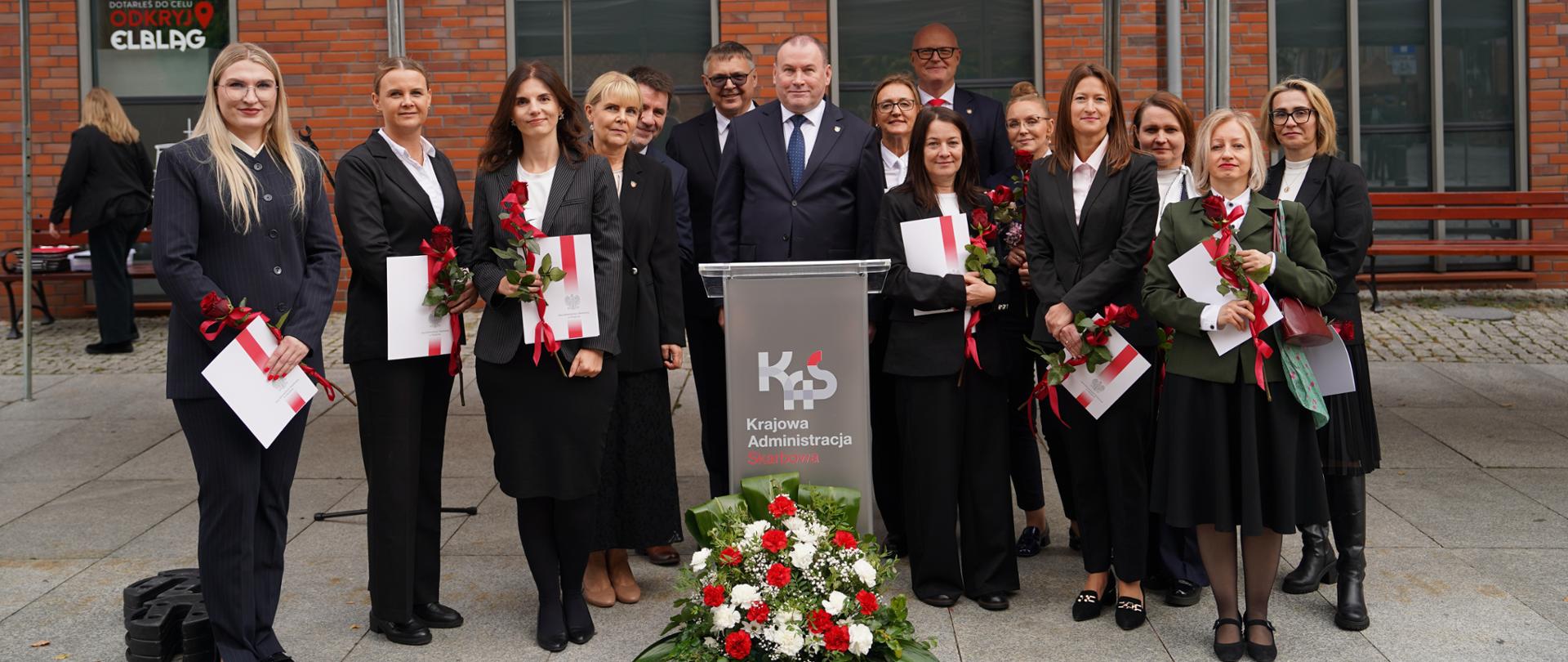 Grupa osób w eleganckich strojach pozuje do wspólnego zdjęcia pod białym namiotem z napisem „Krajowa Administracja Skarbowa”. W centralnej części stoją mężczyźni za mównicą, a wokół niego znajdują się kobiety trzymające dyplomy oraz czerwone róże. W tle widoczny jest budynek z czerwonej cegły.