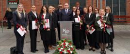 Grupa osób w eleganckich strojach pozuje do wspólnego zdjęcia pod białym namiotem z napisem „Krajowa Administracja Skarbowa”. W centralnej części stoją mężczyźni za mównicą, a wokół niego znajdują się kobiety trzymające dyplomy oraz czerwone róże. W tle widoczny jest budynek z czerwonej cegły.