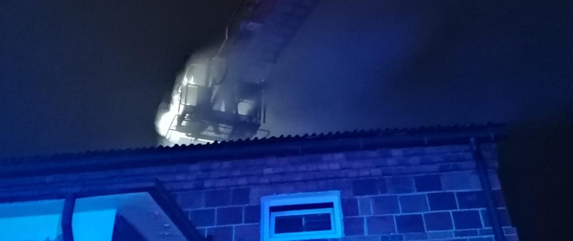 Zdjęcie w porze nocnej. Widać budynek w którym trwał pożar sadzy w kominie. Przed budynkiem pojazd straży pożarnej, podnośnik hydrauliczny.
