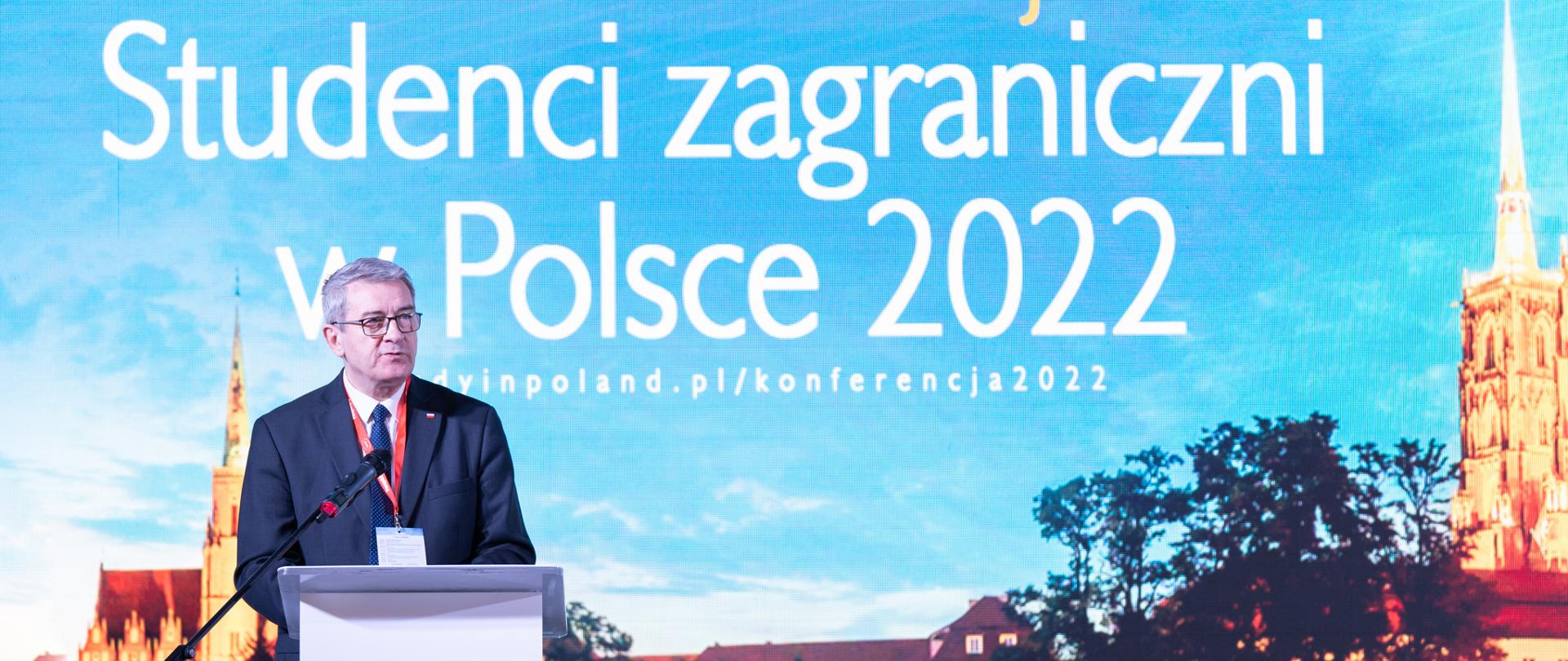 Minister Murdzek przemawia do mikrofonu, za nim wielki plakat z napisem „XV Konferencja - Studenci zagraniczni w Polsce 2022”