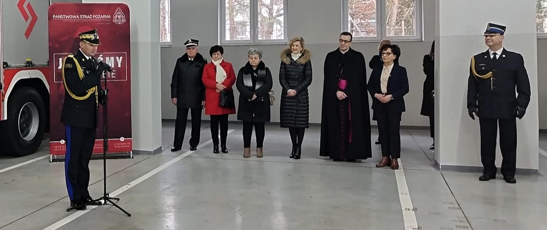 Zastępcą komendanta głównego PSP przemawiający przez mikrofon. W tle pozostali uczestnicy uroczystości. 